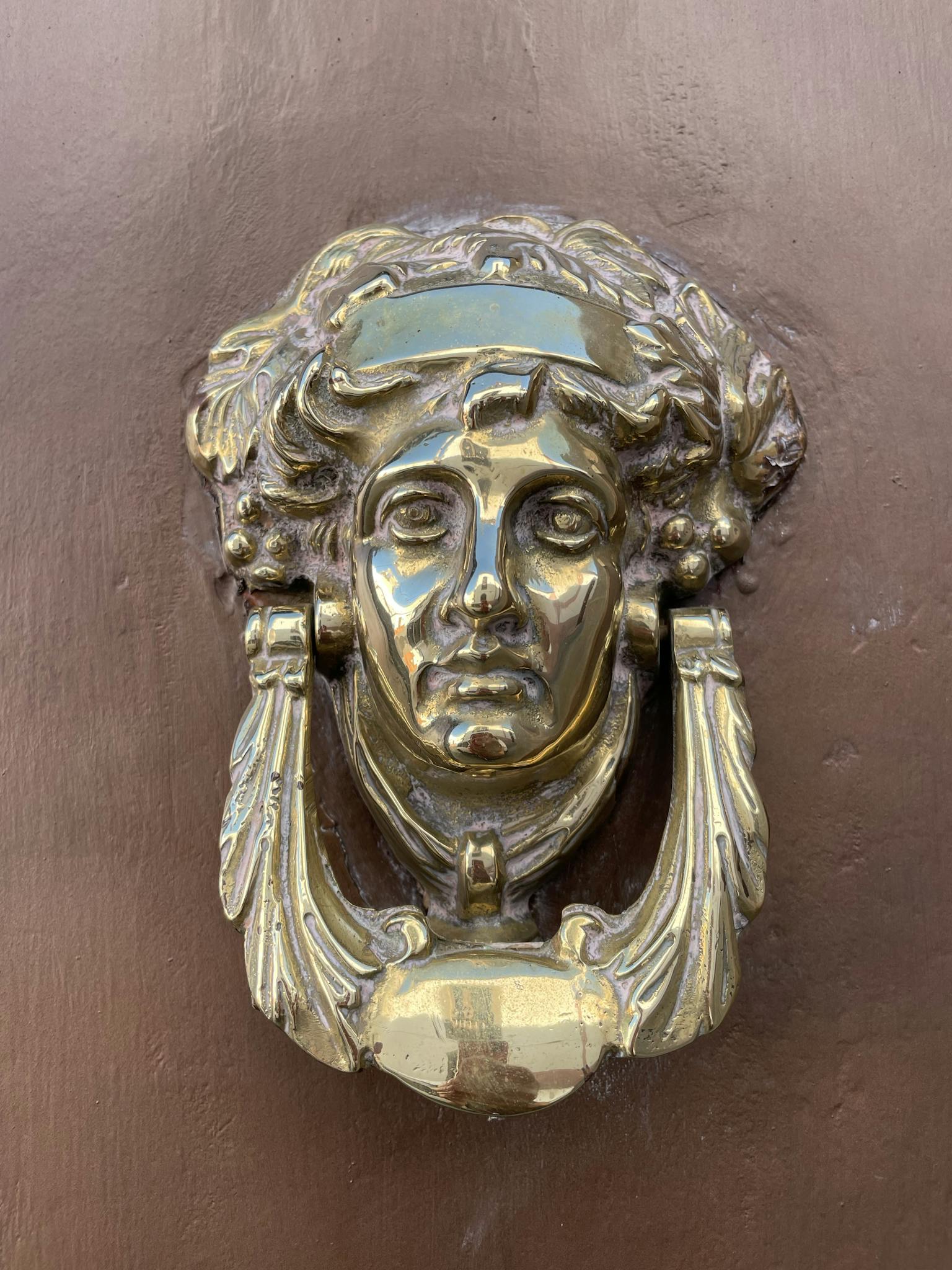 Close-up of a vintage brass doorknocker in the shape of a classical face on a wooden door.